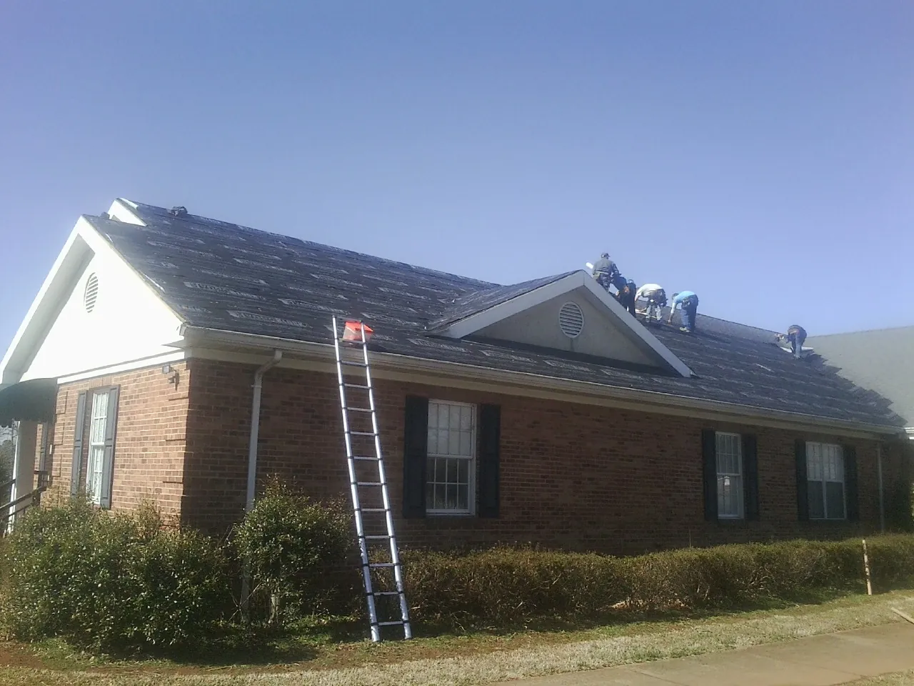 Crew tearing shingles off a brick ranch roof with a ladder set — Metal Roof Repair work by our crew in Kamrar