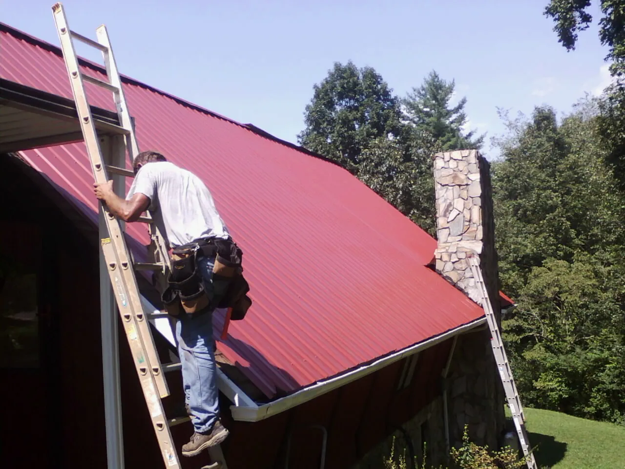 Roofer on a ladder beside a red metal roof and stone chimney — Soffit & Fascia Repair by Roofing Brothers in Kamrar